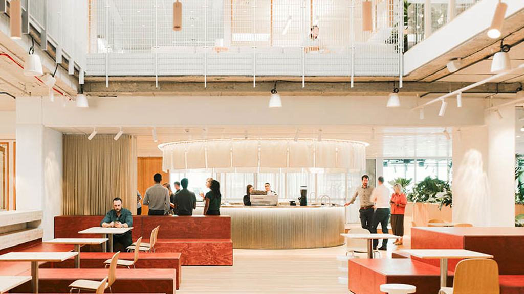 Workplace reception area with large central concierge, tables, chairs, sofas and plants set within warm tones.